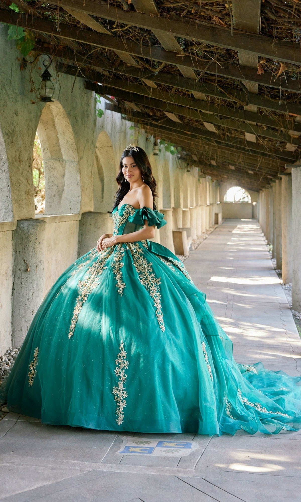 A young woman in the Quinceanera Dress JT1504J By Juliet, a teal ball gown adorned with gold embroidery, stands in a sunlit arched walkway, looking at the camera. The elegant dress features a glitter tulle skirt and a fitted corset bodice, adding sophistication to the radiant scene.